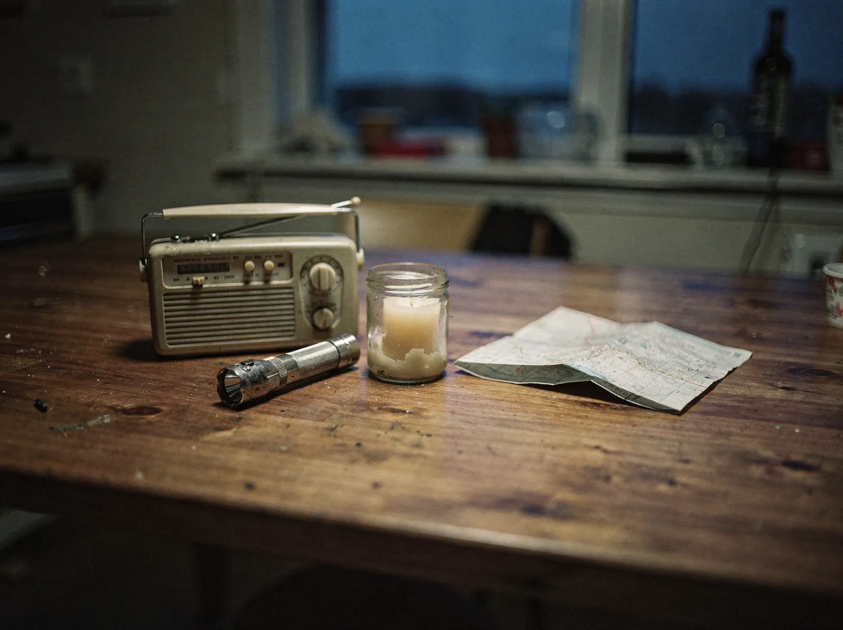 Küchentisch in der Dämmerung mit Kurbelradio, ausgeschalteter Taschenlampe, Kerze im Glas und einer gefalteten Karte