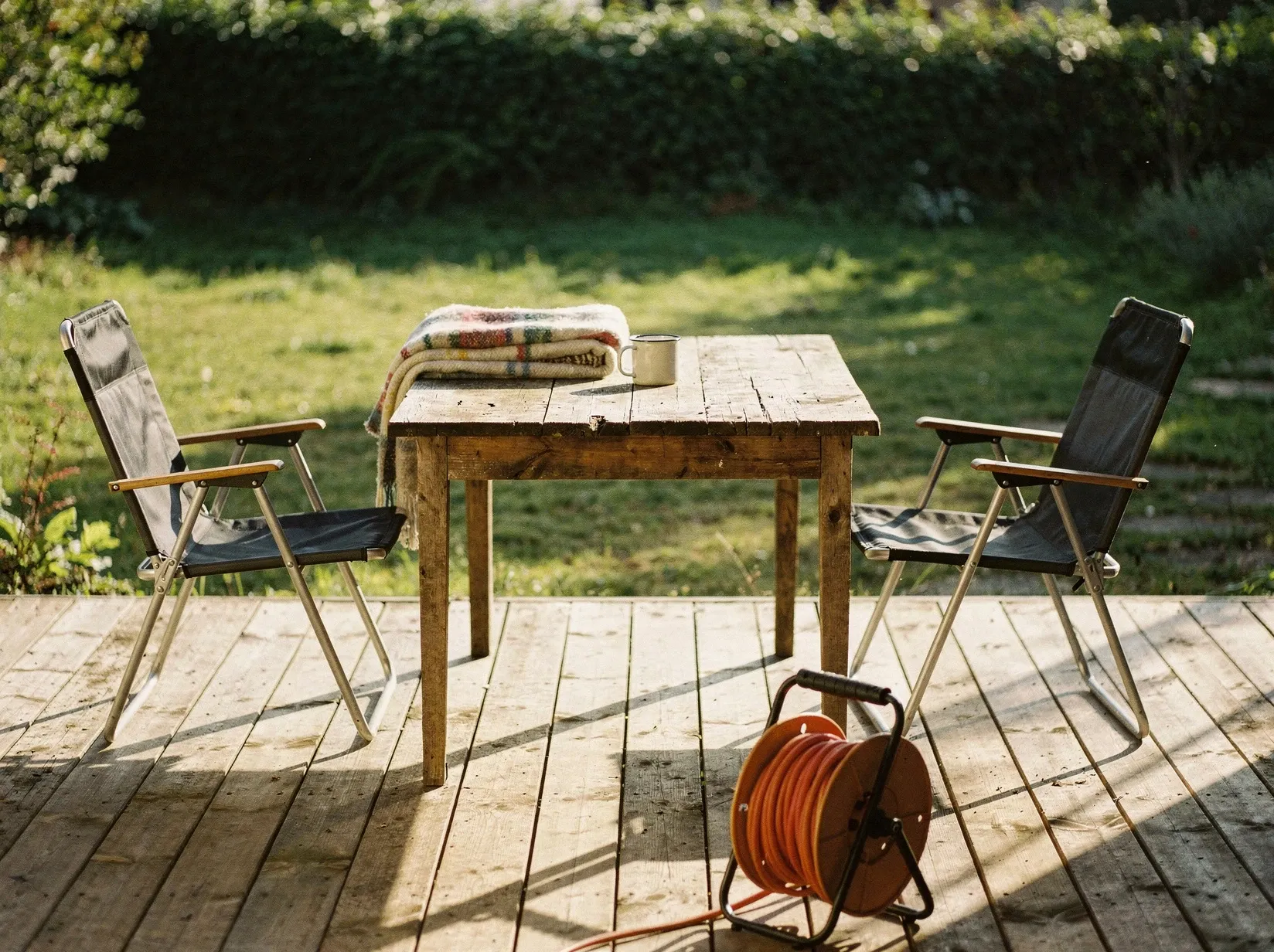 Sonnige Holzterrasse am Nachmittag mit Holztisch, zwei Campingstühlen und einer aufgerollten Kabeltrommel, im Hintergrund ein grüner Garten