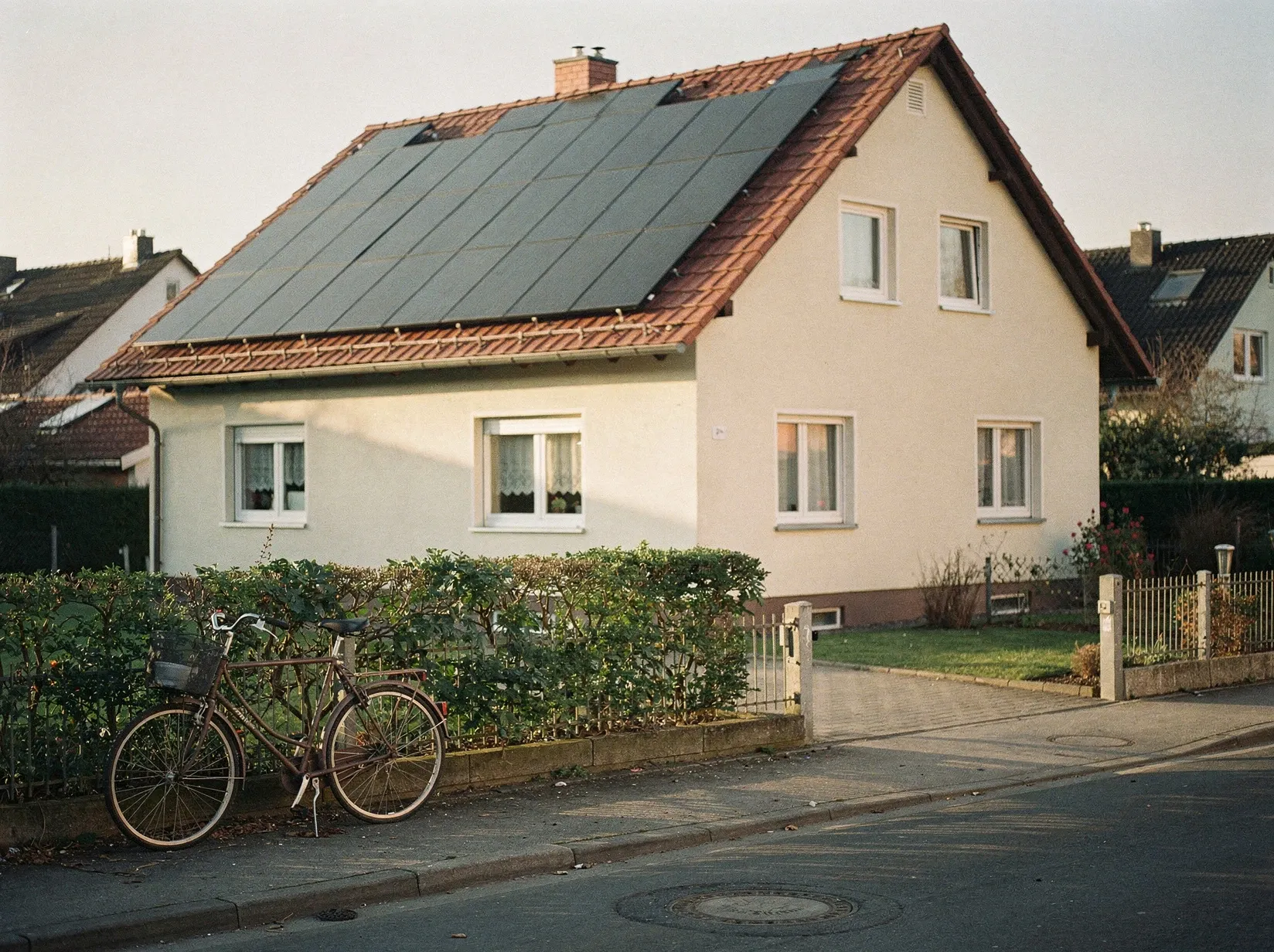 Wohnhaus in einer Vorortstraße mit Photovoltaik-Modulen auf dem Dach im Spätnachmittagslicht, davor Hecke und ein abgestelltes Fahrrad
