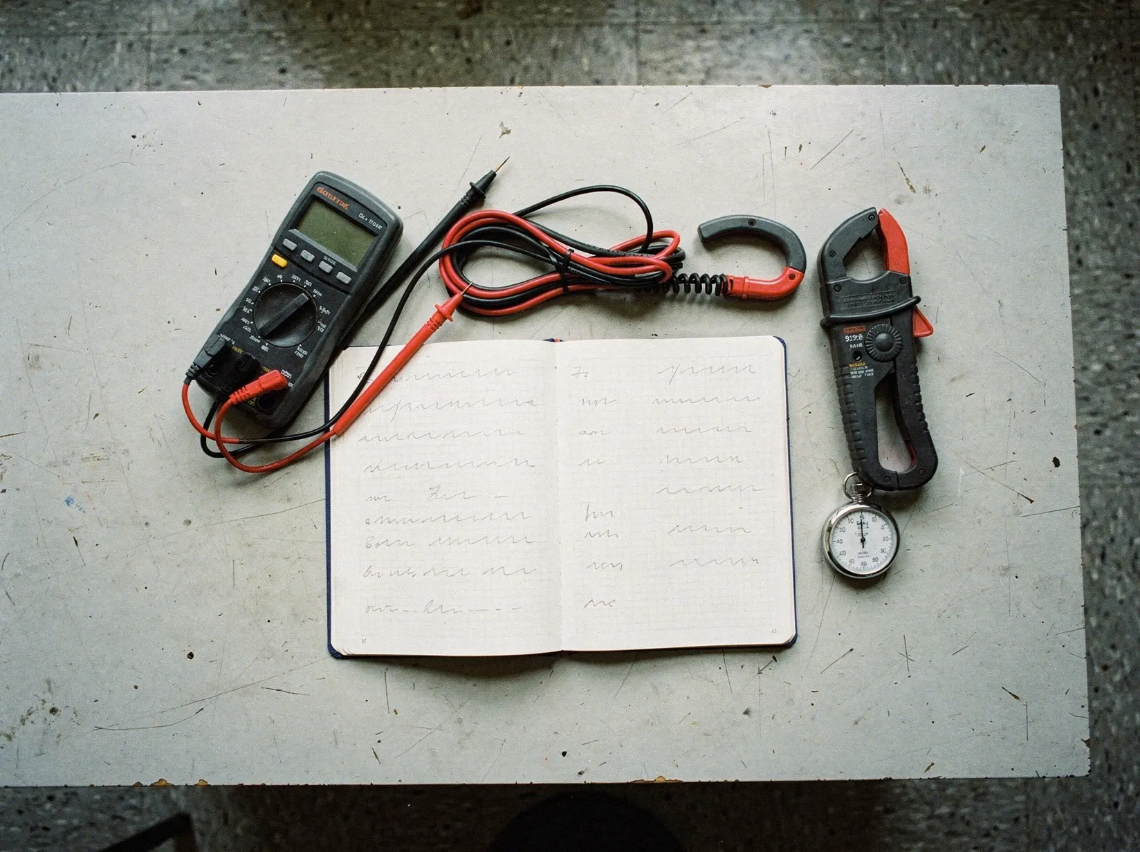 Werkbank mit ausgeschaltetem Multimeter, Zangenamperemeter, einem aufgeschlagenen Notizbuch mit Messreihen und einer mechanischen Stoppuhr