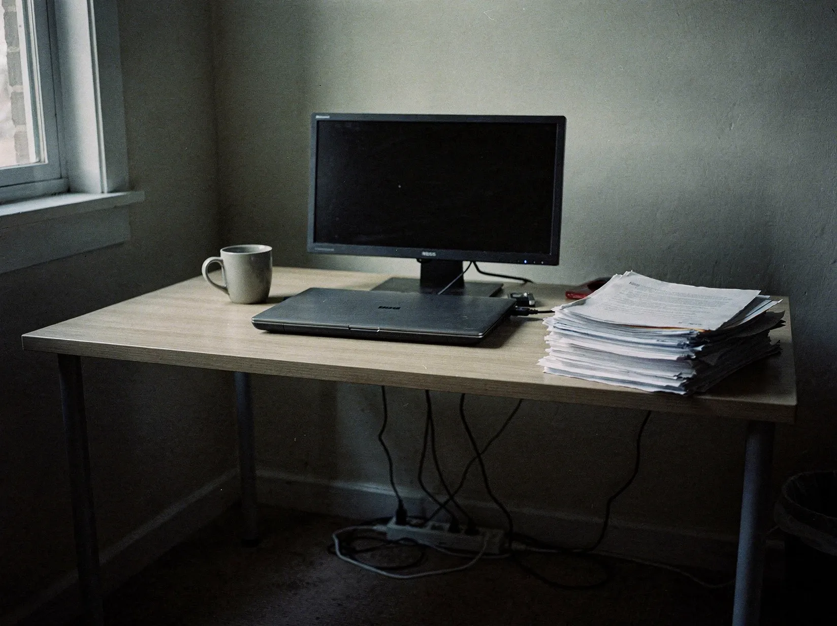 Schreibtisch im Homeoffice in der Dämmerung mit ausgeschaltetem Monitor, geschlossenem Laptop und einer Steckdosenleiste am Boden