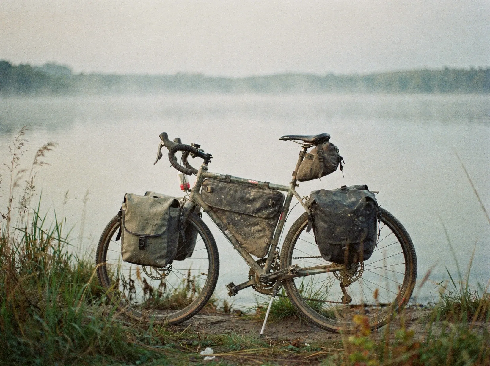 Reiserad mit Packtaschen am Ufer eines Sees im Morgennebel, Schilf im Vordergrund