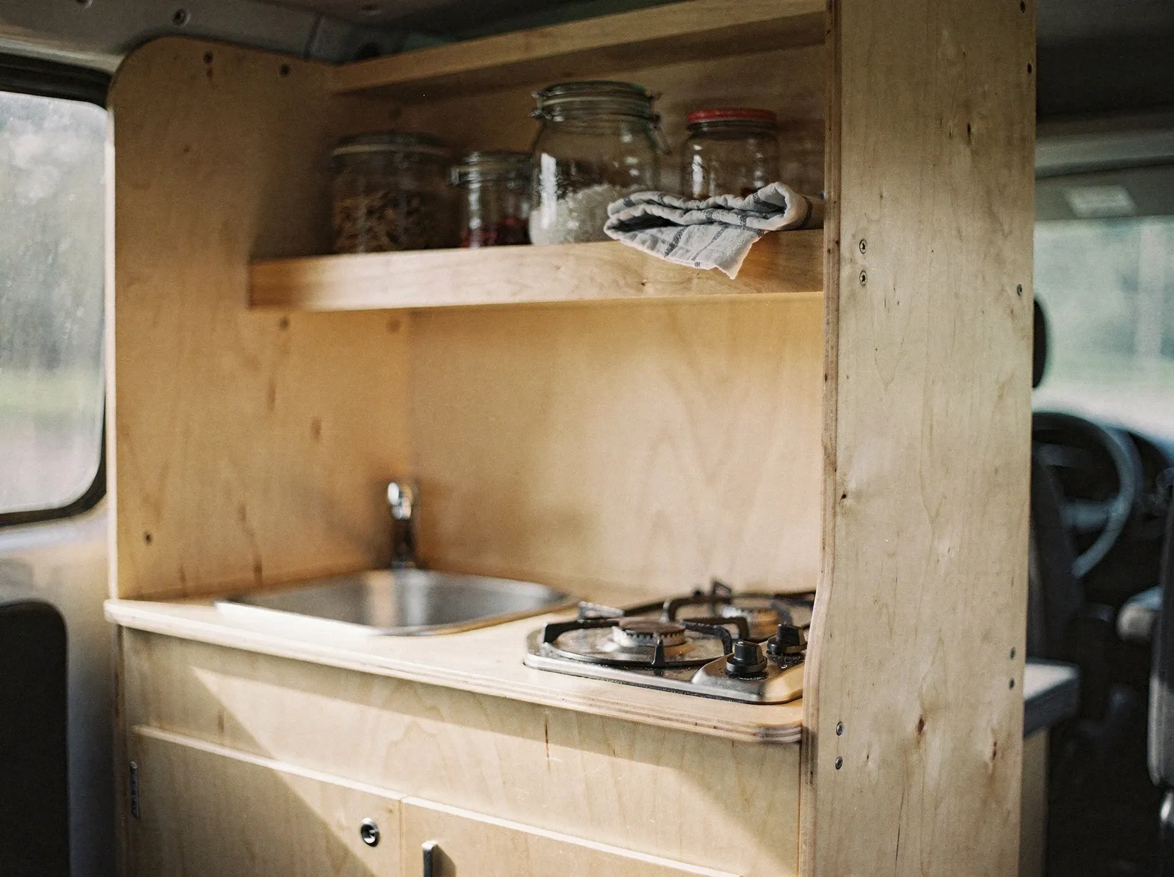 Selbstausgebaute Camper-Küche aus hellem Sperrholz mit kleiner Spüle und Gaskocher, Nachmittagslicht durch das Seitenfenster