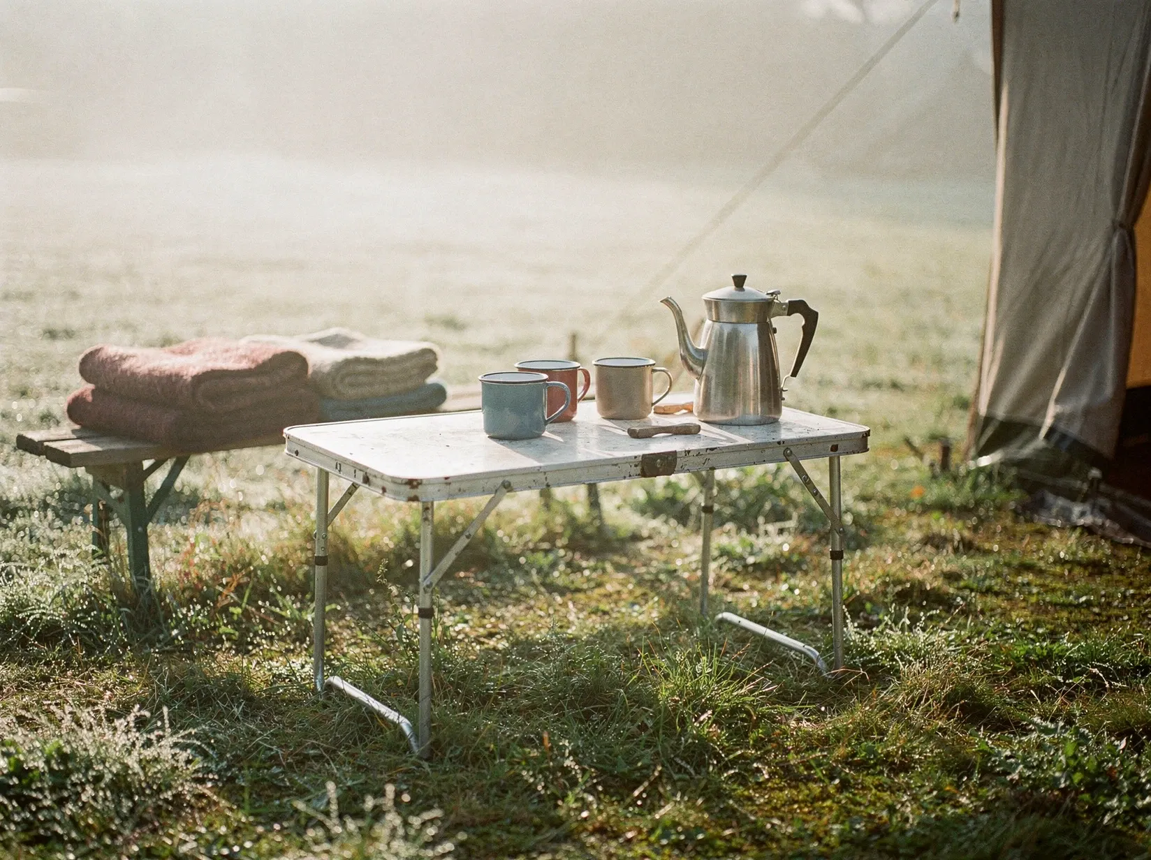 Campingtisch auf einer Wiese im Morgendunst, drei Emaillebecher und eine Kanne, daneben ein Zelt