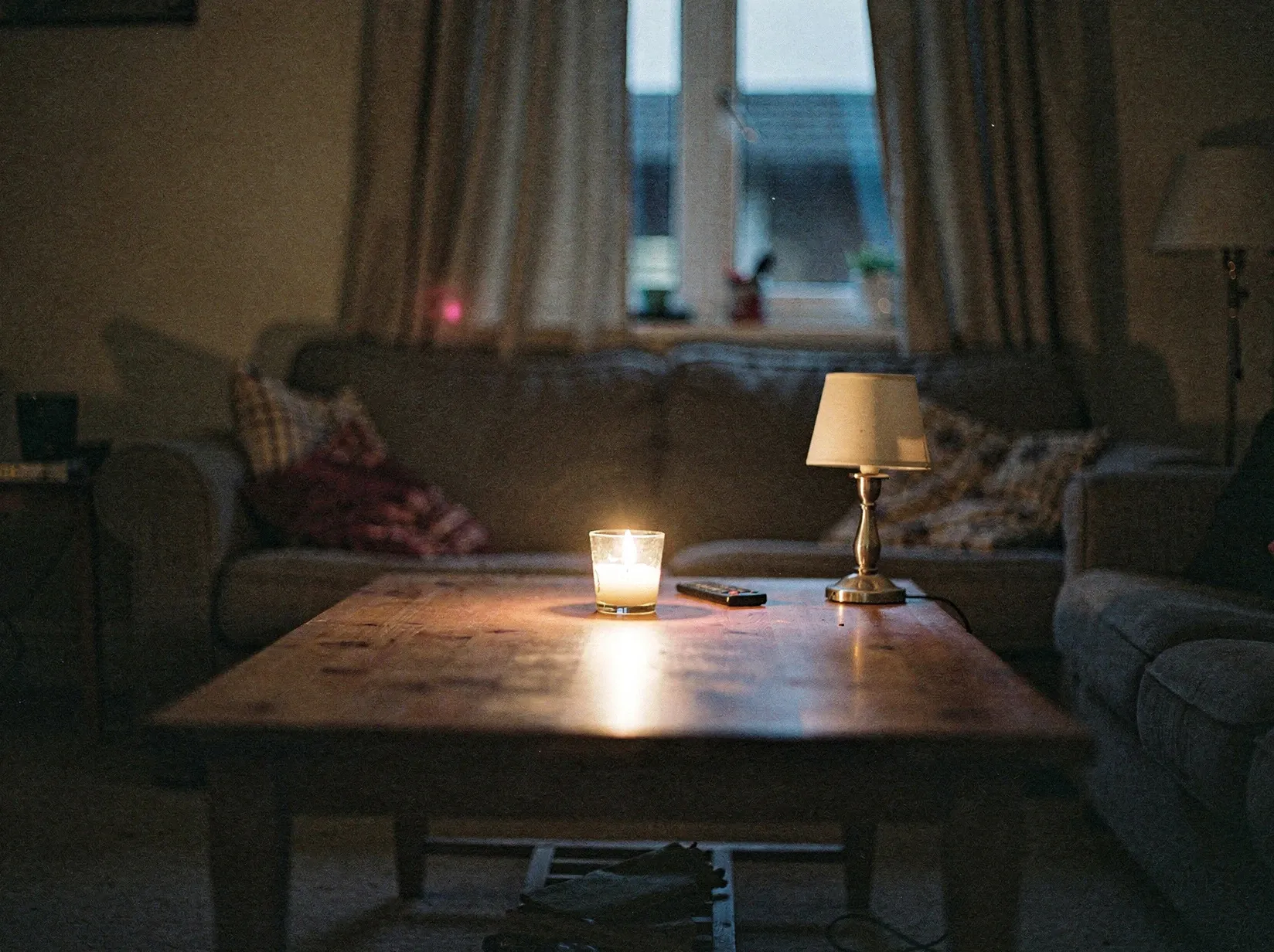 Wohnzimmer bei Stromausfall in der Dämmerung, eine brennende Kerze auf dem Couchtisch neben einer ausgeschalteten Lampe
