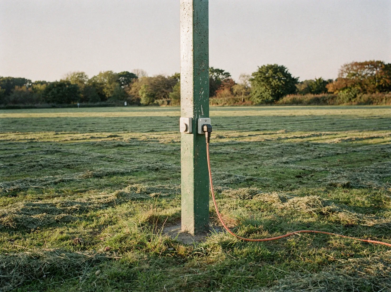 Stromsäule auf einer Campingplatzwiese, ein orangefarbenes Verlängerungskabel steckt in der Steckdose und führt über das Gras